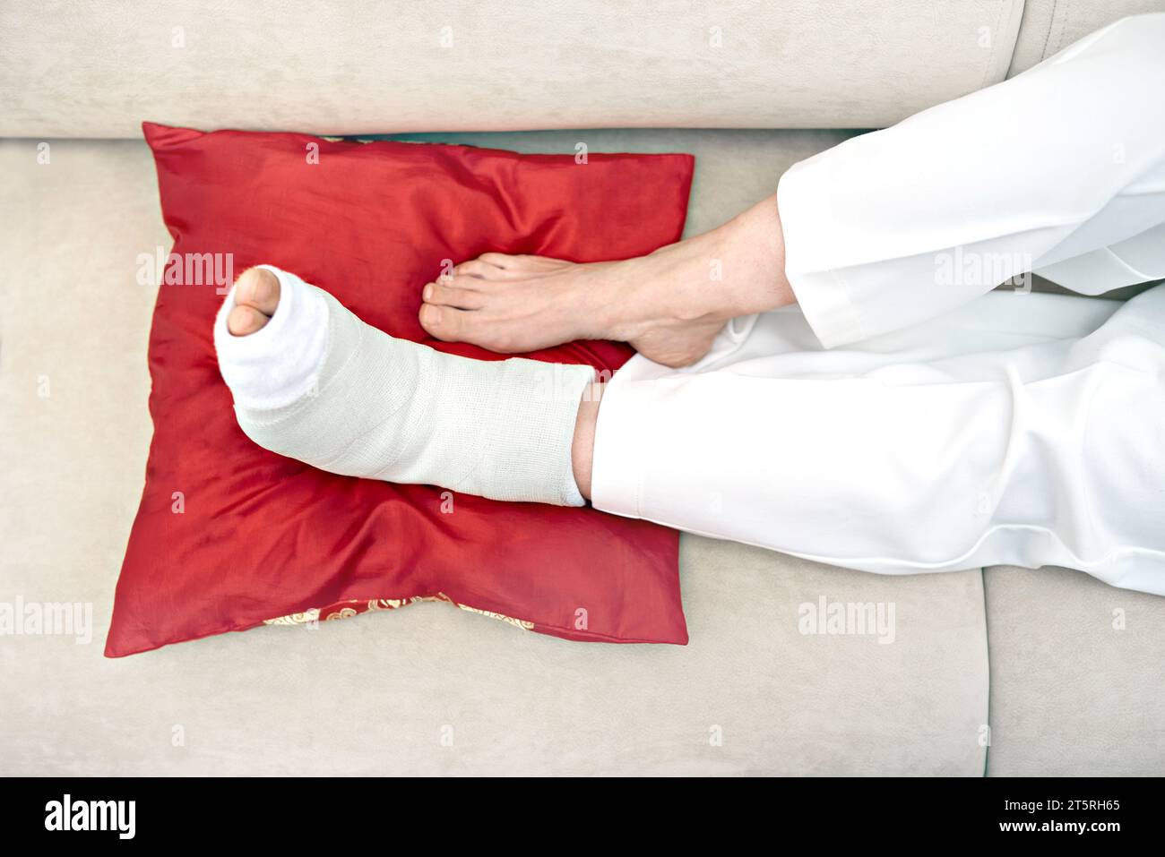 Female broken foot in white plaster cast lying on a red pillow resting. Injury, trauma, recovery, rehabilitation of people concept . Therapy at home f Stock Photo