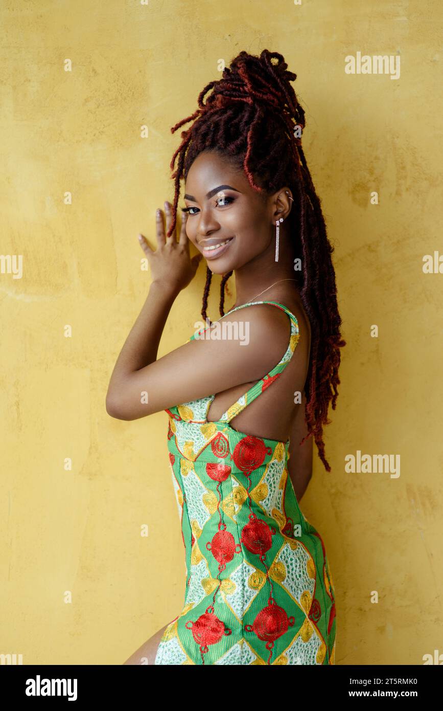 Semi-profile waist up portrait of the smiling young African woman with flawless skin and make-up, wearing a bright dress, looking at the camera and sm Stock Photo