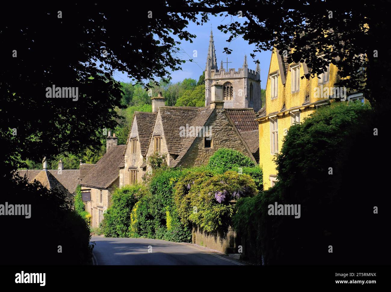 Castle Combe: St Andrew’s Church, Cotswold stone cottages and wisteria in bloom in Castle Combe village, Cotswolds, Wiltshire, England, UK Stock Photo