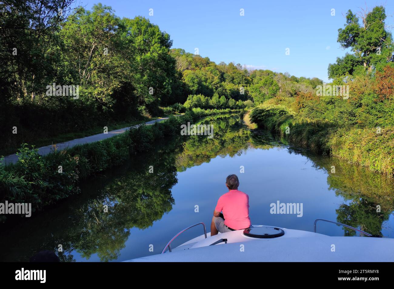Canal du Nivernais: Cruising along Nivernais Canal with older man on bow of boat and mirror reflections of trees in Burgundy, France Stock Photo