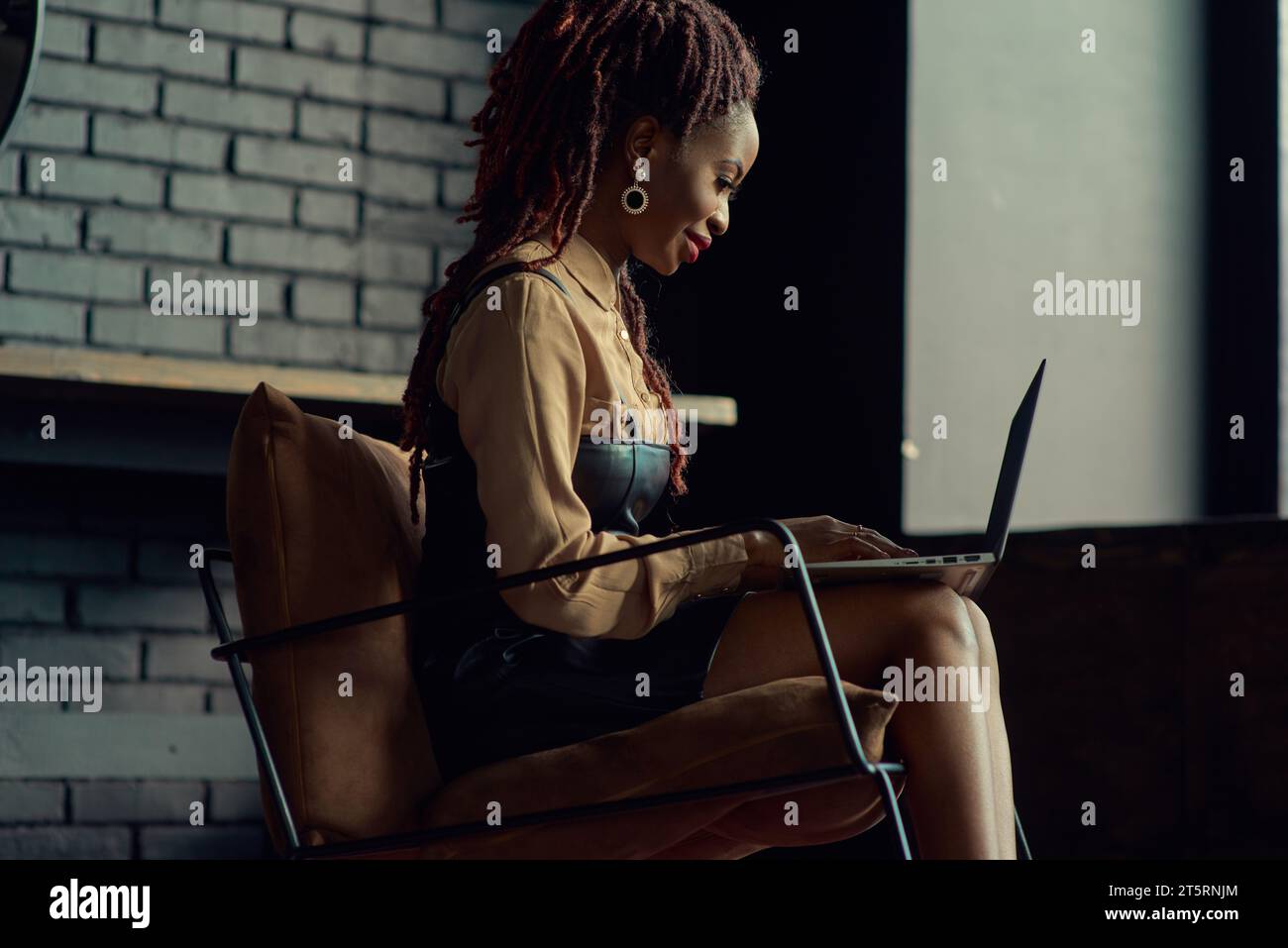 Black woman sitting in modern office, stylish loft apartment and typing on a laptop. Entrepreneur, fleelancer doing remote work. Business woman, compu Stock Photo