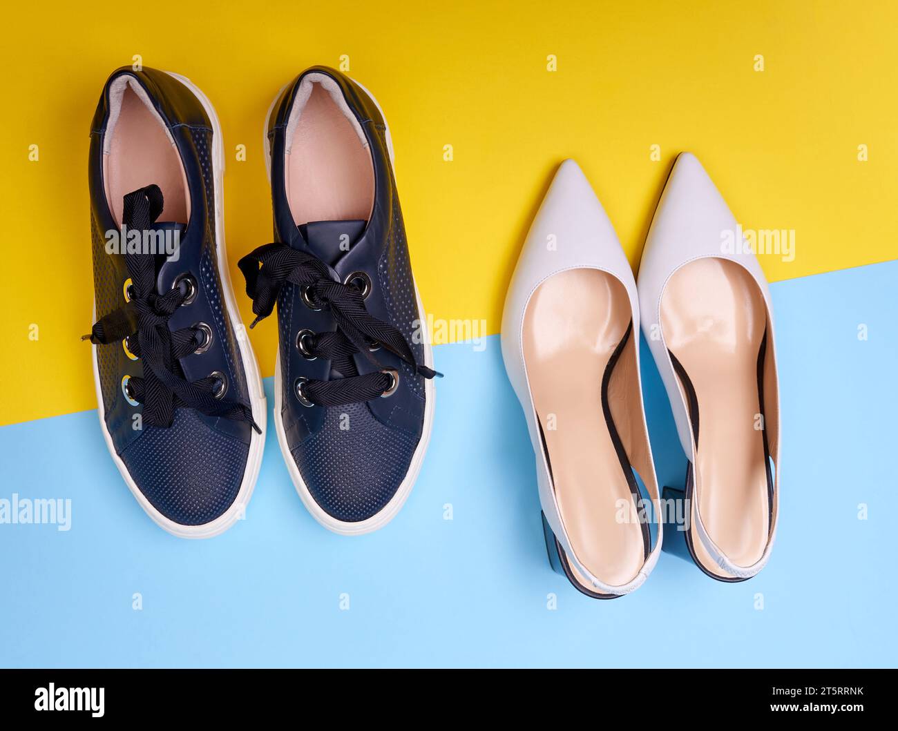 Two pairs of female modern shoes: open-back white pumps with black flare heels and casual navy blue sneakers on a blue-yellow background. Top view, cl Stock Photo