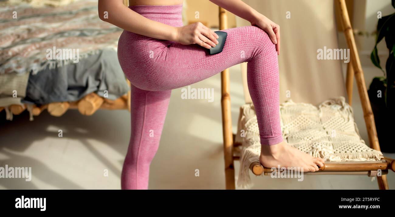 Woman in pink leggings using black comb edge gua sha tool on her leg to relieve muscular tensions. Lower body shot. Physical therapy and wellness. Tra Stock Photo