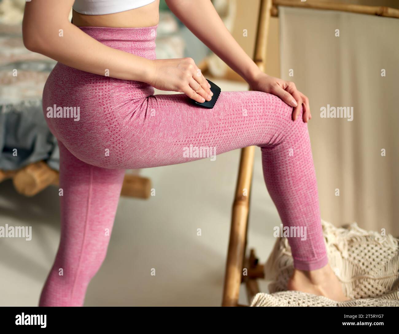 A lady dressed in pink leggings is using a black-edged Gua Sha comb on her leg to relieve muscle tension. Rehabilitation and wellness concept,   pract Stock Photo