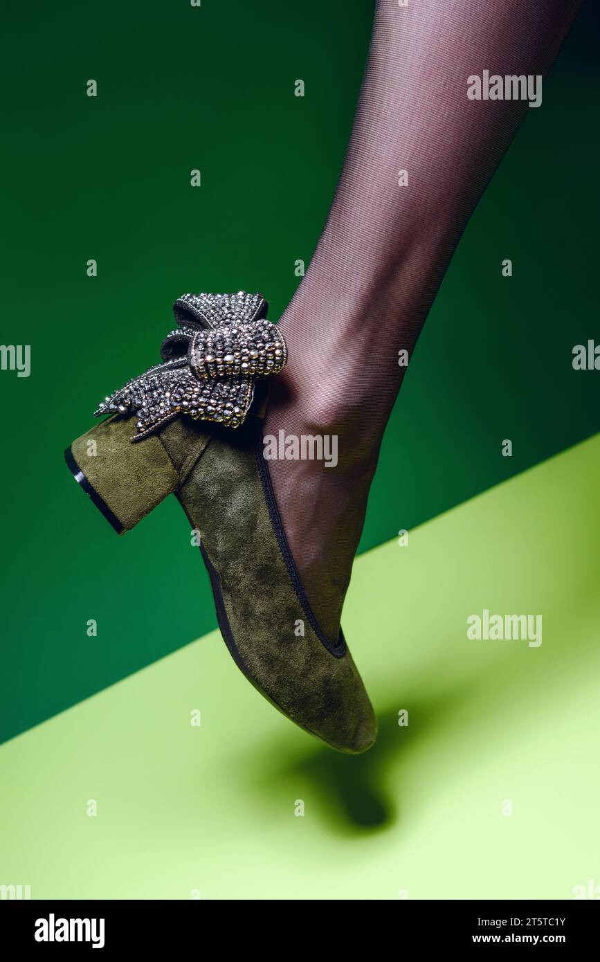 Elegant block-heeled green suede shoe decorated with a shiny silver bow on a slender female leg in black nylon tights on a two-tone green background. Stock Photo