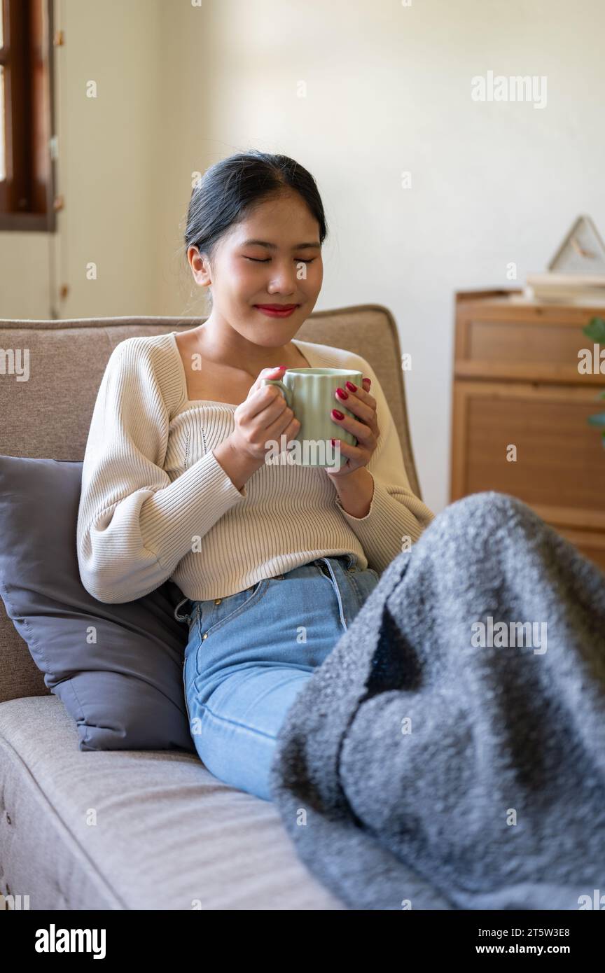 A happy, beautiful Asian woman in casual clothes is smelling her coffee, enjoying her morning coffee on a couch in her minimalist living room. Domesti Stock Photo