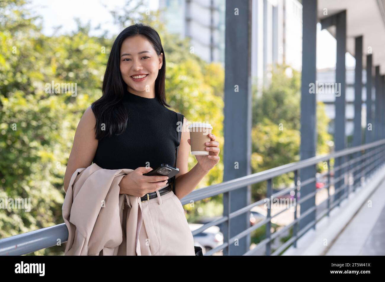 An attractive, gorgeous millennial Asian businesswoman is on a skywalk in the city, smiling at the camera, and holding a takeaway coffee cup and her p Stock Photo