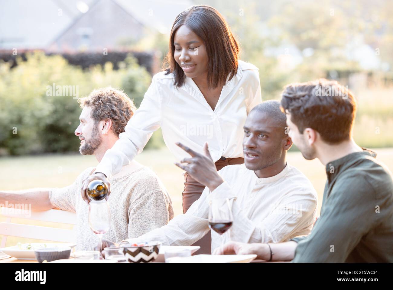 Amidst the ambient sounds of laughter and clinking glasses, a diverse group of millennial friends comes together in a garden setting, celebrating their bond over a sumptuous dinner. The heart of the gathering is the gracious hostess - the black lady of the house. With a wine bottle in hand, she fills a guest's glass, her gesture embodying the spirit of generosity and togetherness that defines the evening. Cheers to Friendship: Hostess Pours Wine at Multiracial Garden Dinner Party. High quality photo Stock Photo