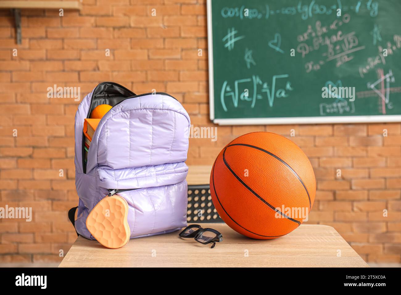 Stylish school backpack with stationery, ball and eyeglasses on desk in classroom Stock Photo