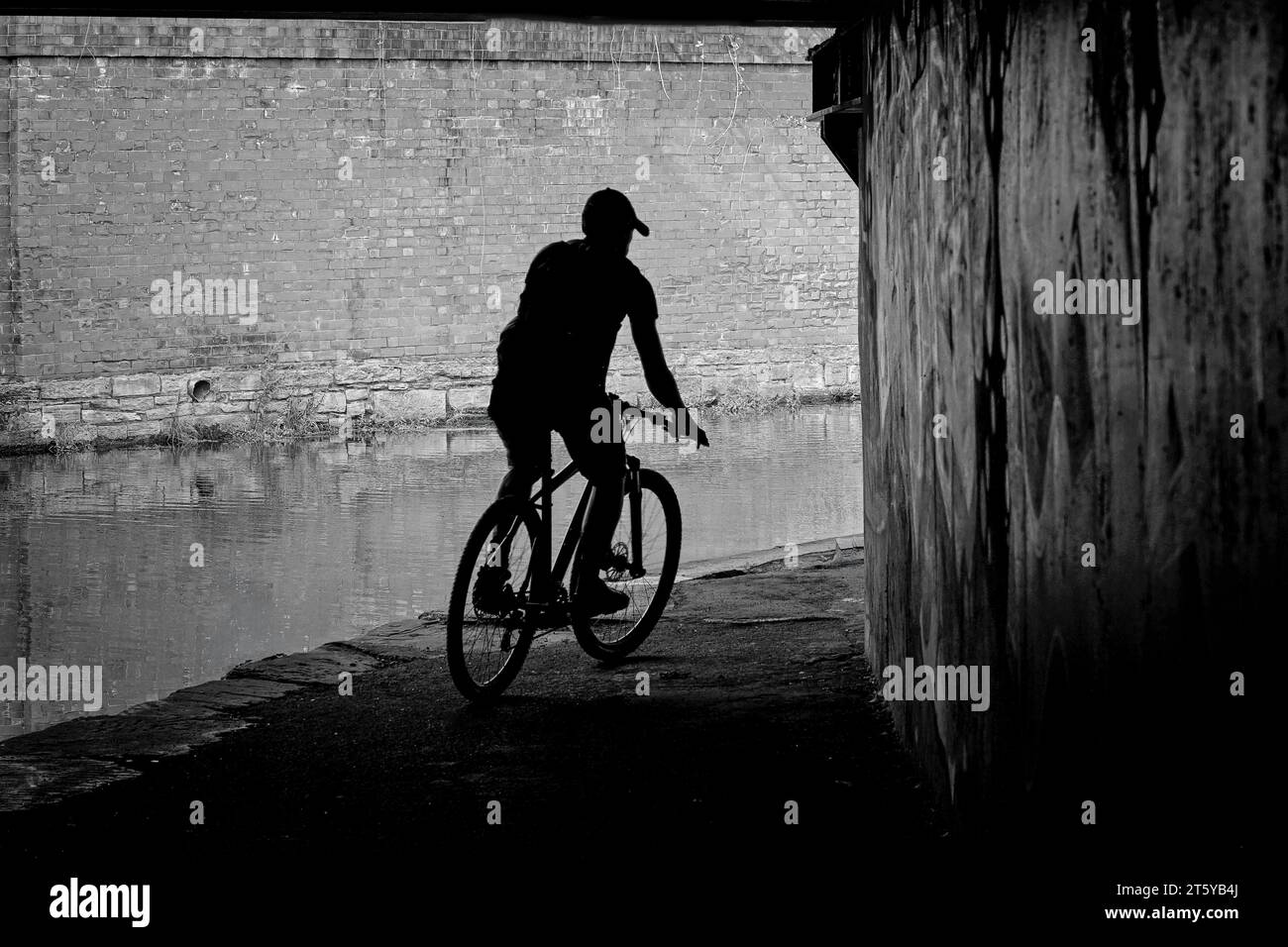 A male cyclist is silhouetted against a brick wall as he rides beneath a bridge along a narrow canal in Huddersfield, Yorkshire, England, UK. Stock Photo