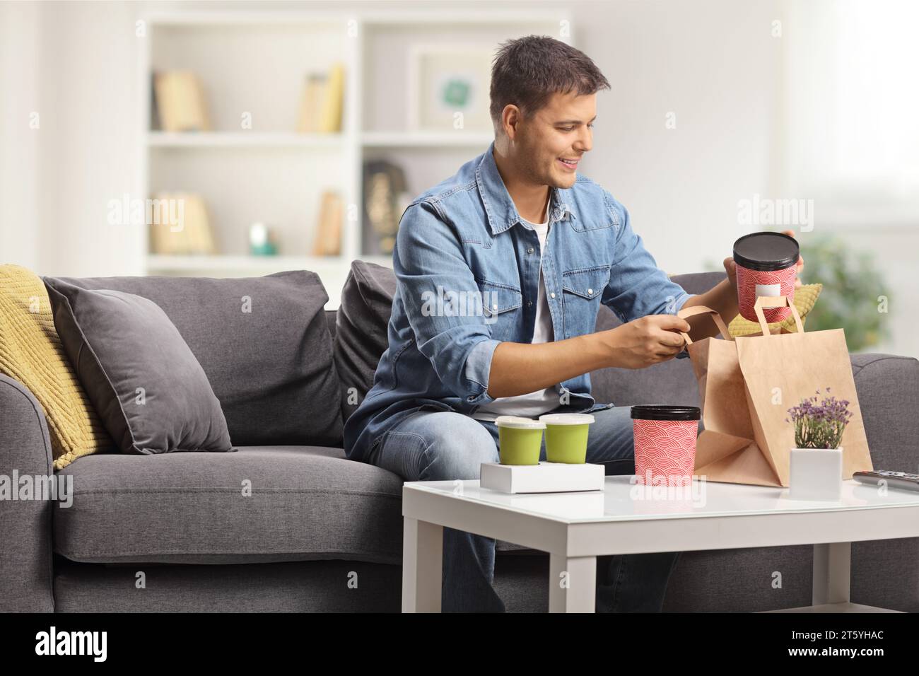 Happy young man sitting on a sofa and opening takeaway food boxes at home Stock Photo