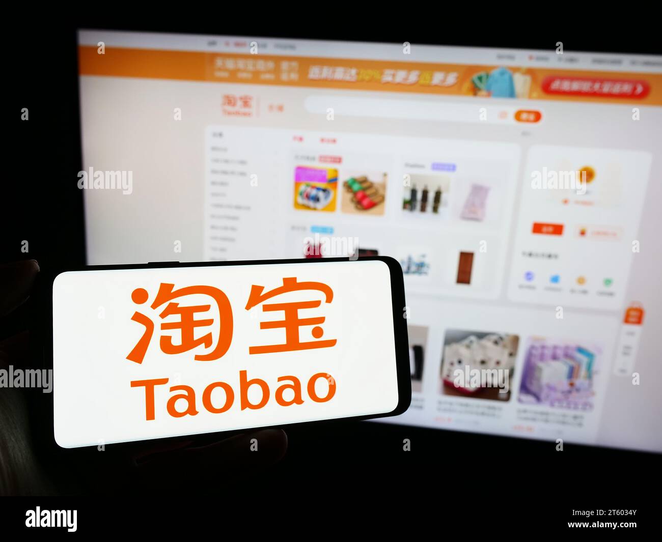 Person holding mobile phone with logo of Chinese online shop Taobao (Alibaba Group) in front of company web page. Focus on phone display. Stock Photo
