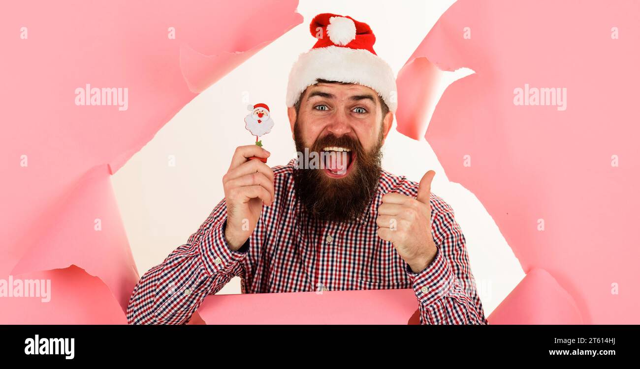 Happy bearded man in Santa hat with little Santa candy lollipop showing thumb up looking through paper hole. Christmas decoration. Holiday poster Stock Photo