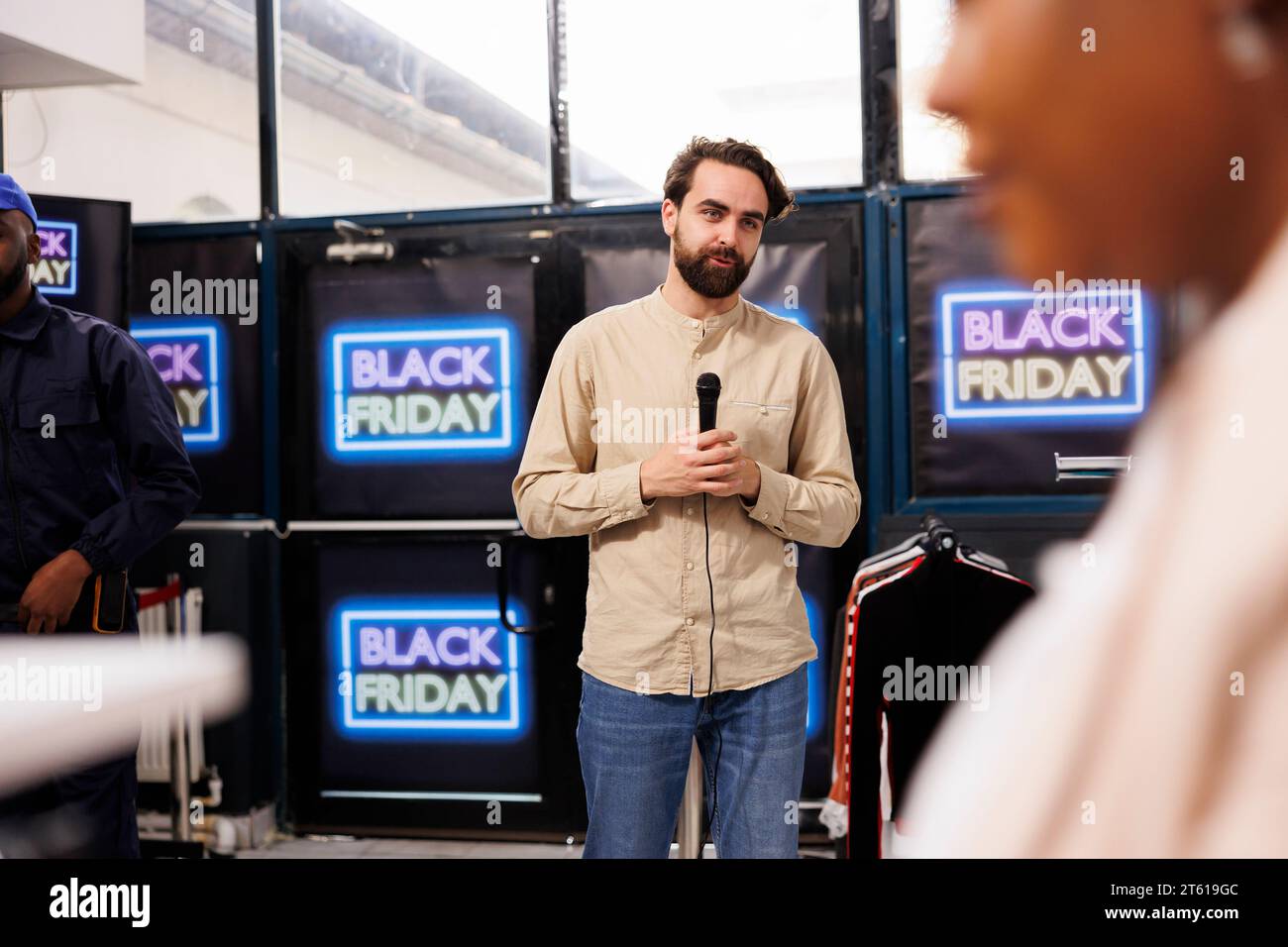 Male tv reporter standing in front of camera with microphone in hands reporting breaking news from Black Friday event. News reporter live broadcasting from crowded shopping mall during seasonal sales Stock Photo