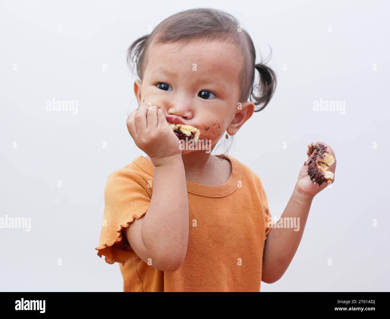 cute little Asian girl eating sweet donut or chocolate bread, mouth covered in chocolate. Stock Photo