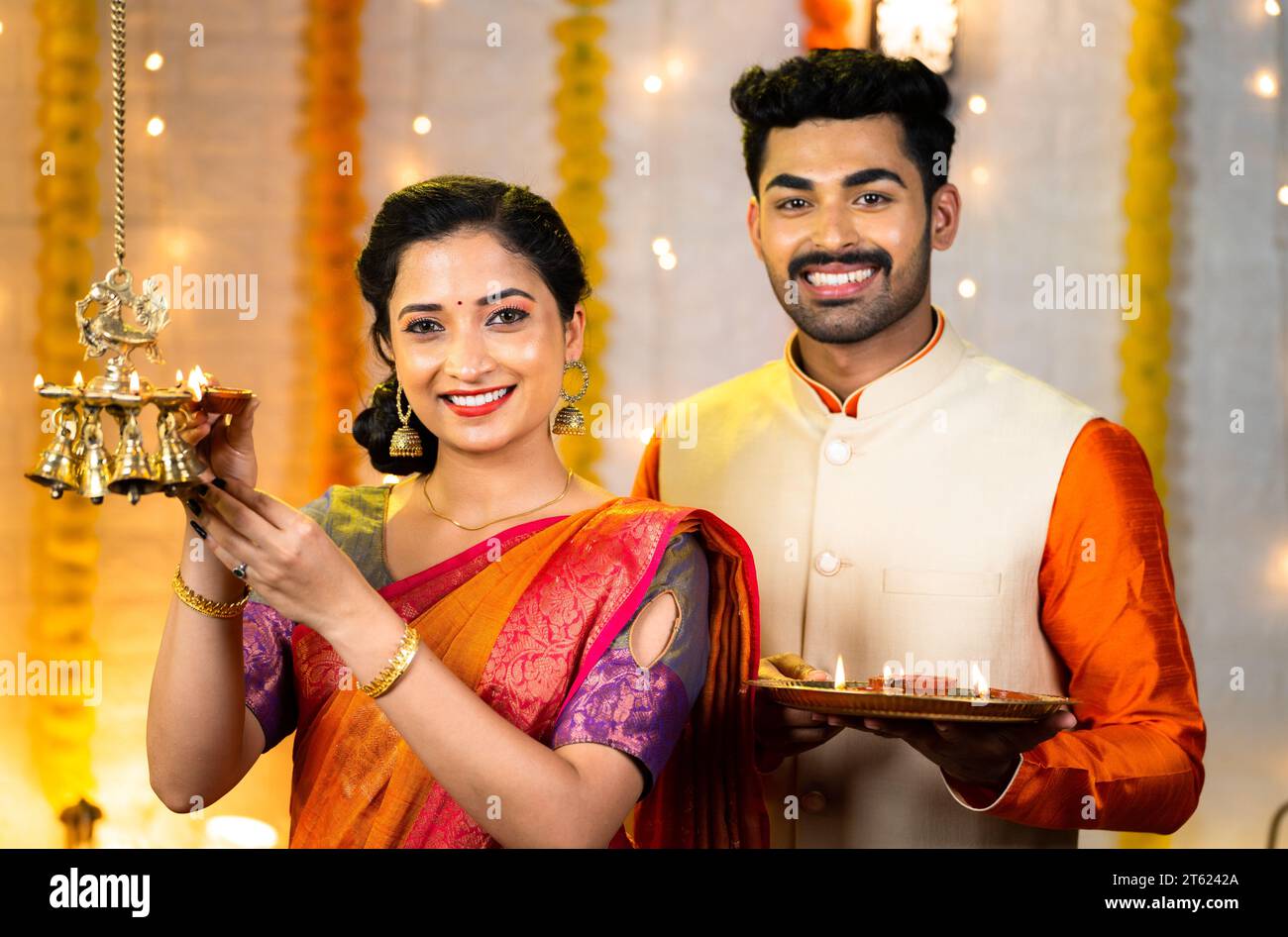Happy indian couple in traditional ethnic wear lighting brass hanging oil lamp by looking at camera at home - concept of diwali celebration, togethern Stock Photo
