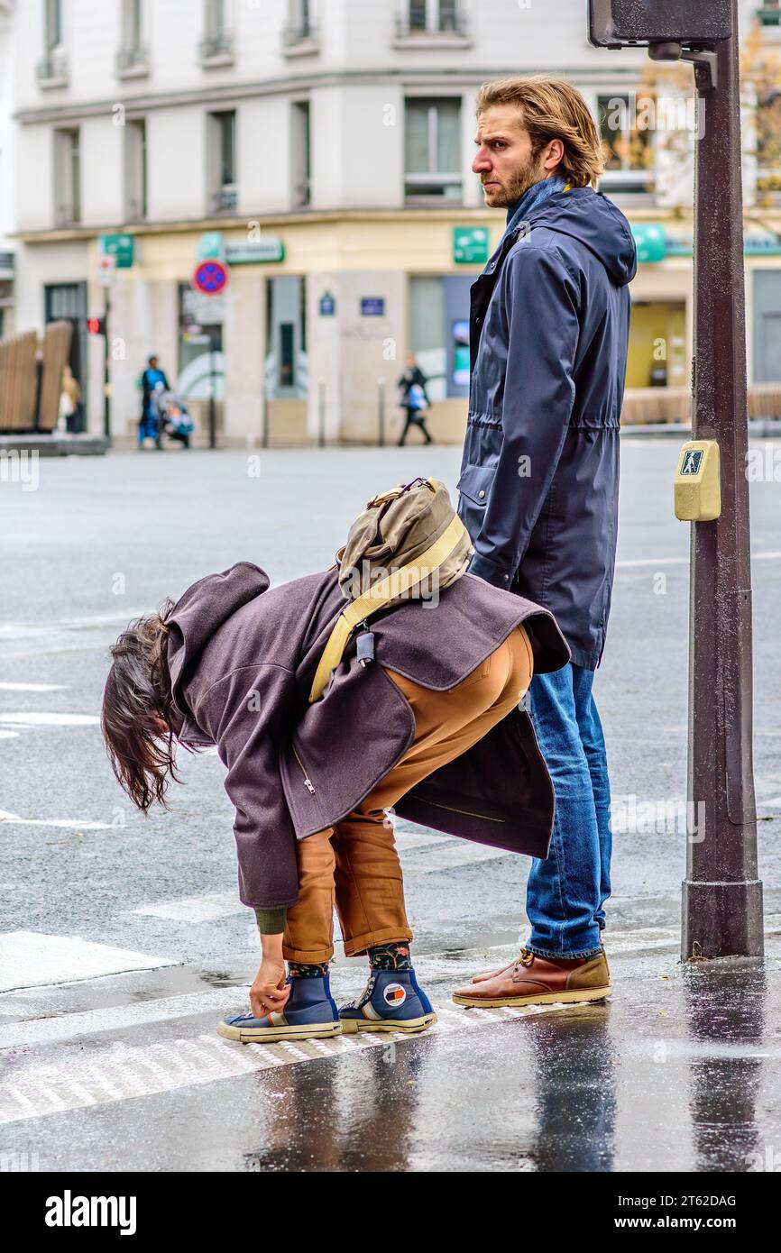 Couple waiting at street crossing with woman retying shoelace - Paris 20, France. Stock Photo