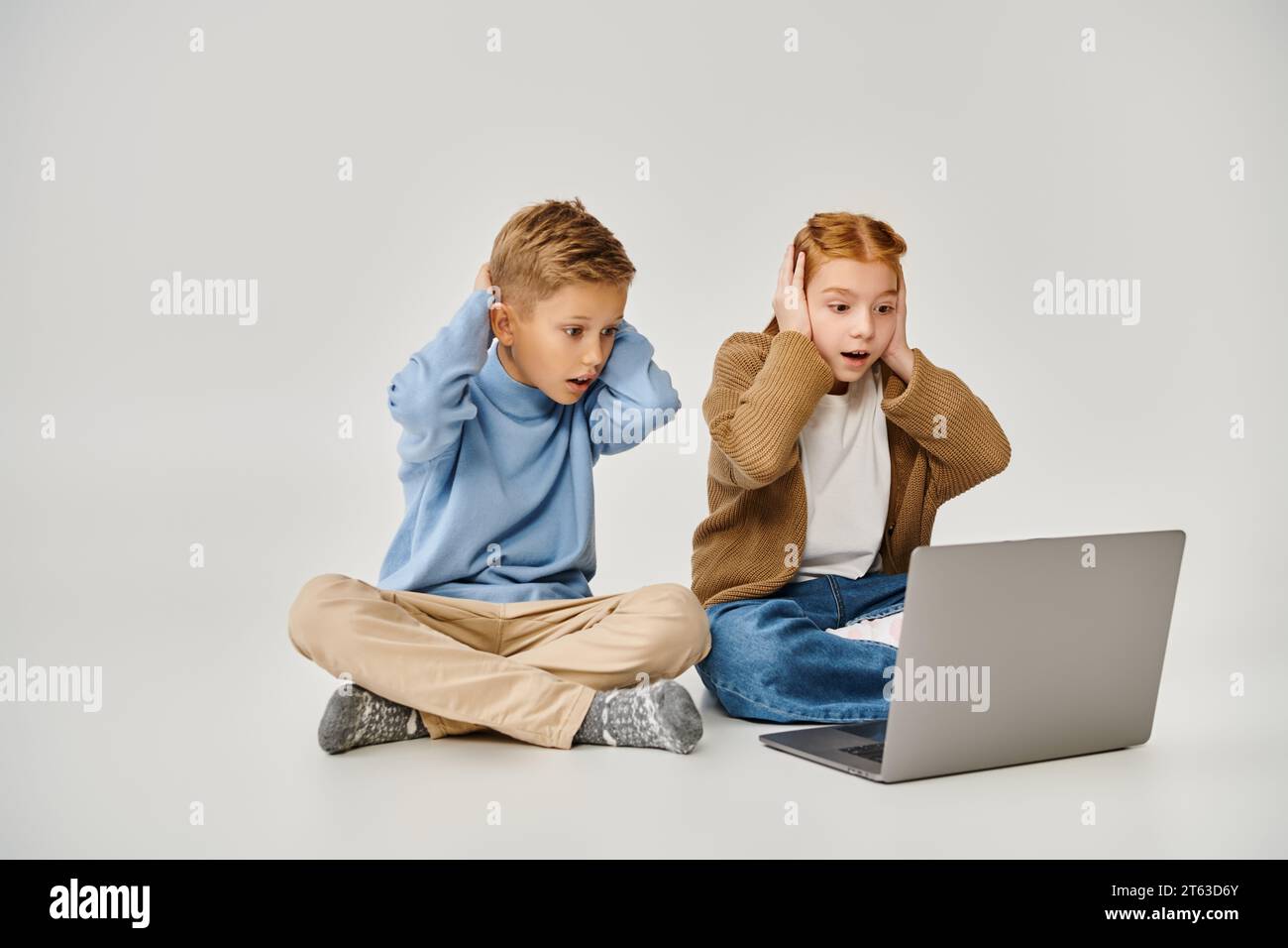 two scared trendy little children looking at laptop and closing ears ...