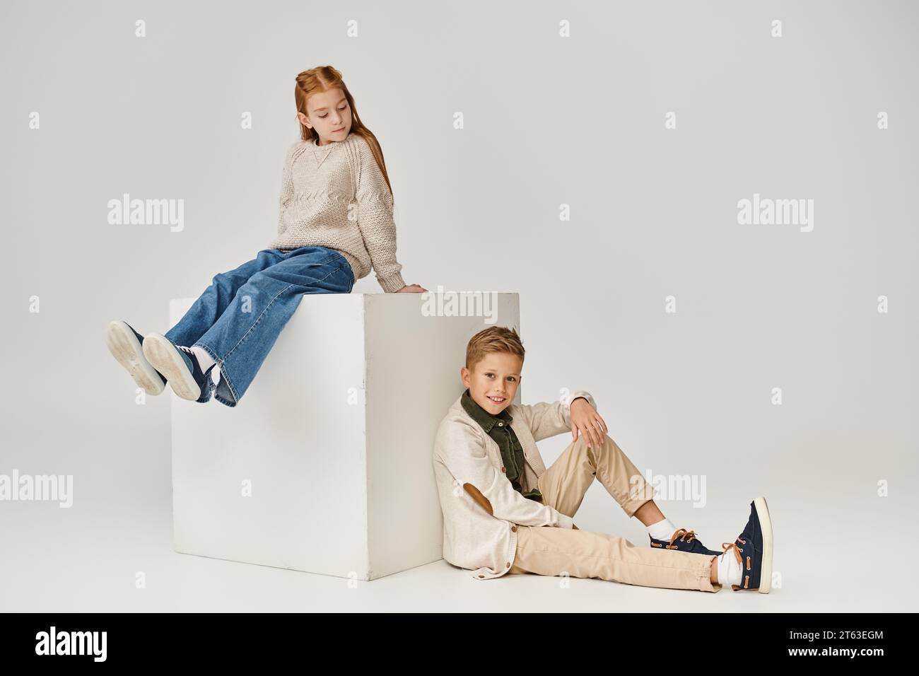 red haired little girl sitting on cube and looking at smiley preteen boy sitting on floor, fashion Stock Photo
