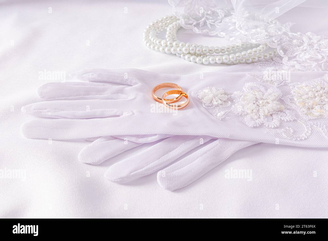 A pair of gold wedding rings lie on luxuriouswedding white gloves. White satin background with pearl beads. Wedding accessories concept Stock Photo