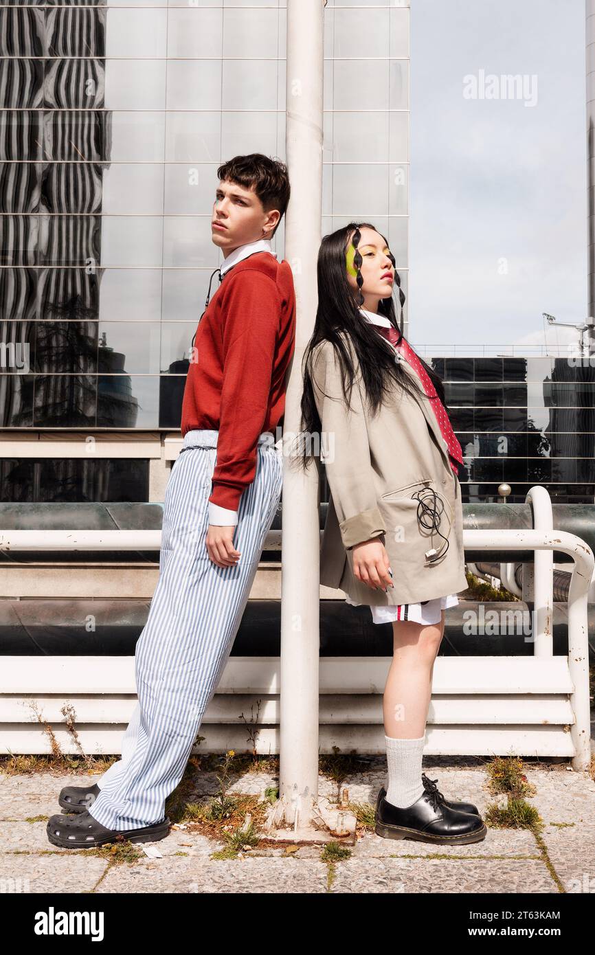 Two young Gen-Z individuals with cables leaning on a lamppost fashionably against a modern building in a work concept. Stock Photo