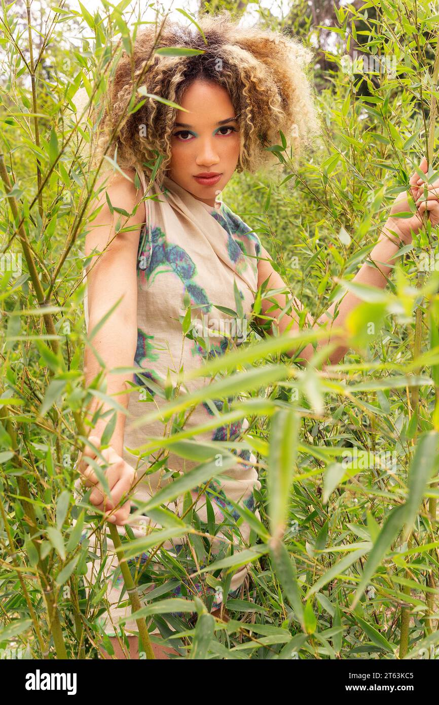 Ethnic young woman with curly blonde hair stands amidst tall green ...