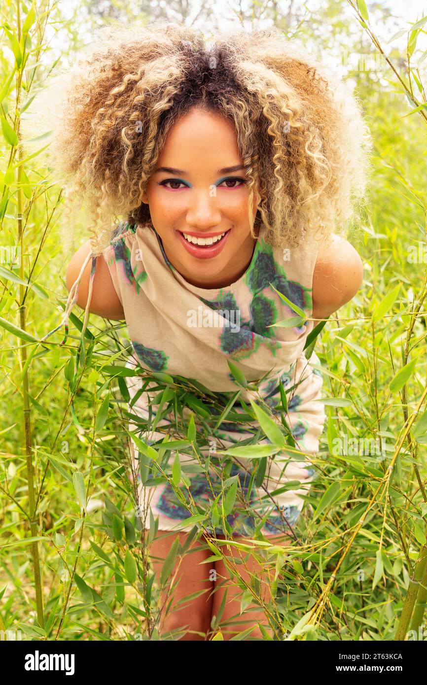 Ethnic young woman with curly blonde hair stands amidst tall green ...