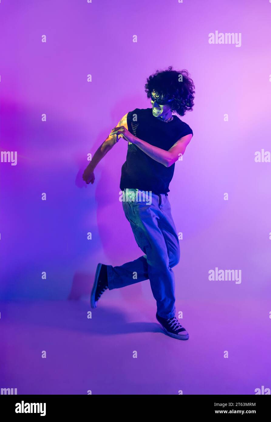 A curly-haired male exudes energy as he dances dynamically against a lilac-lit backdrop, with vivid neon accents highlighting his movement Stock Photo