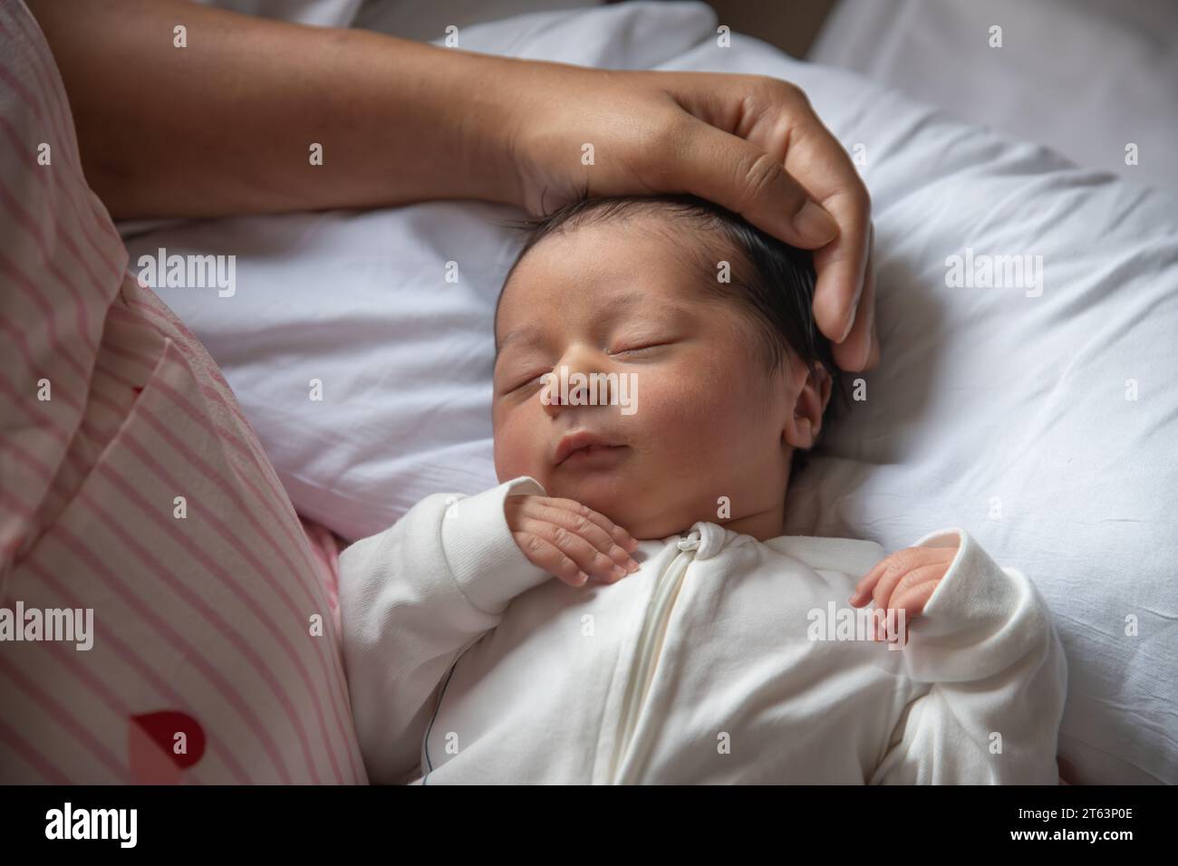 A gentle hand caresses the head of a sleeping newborn baby, dressed in