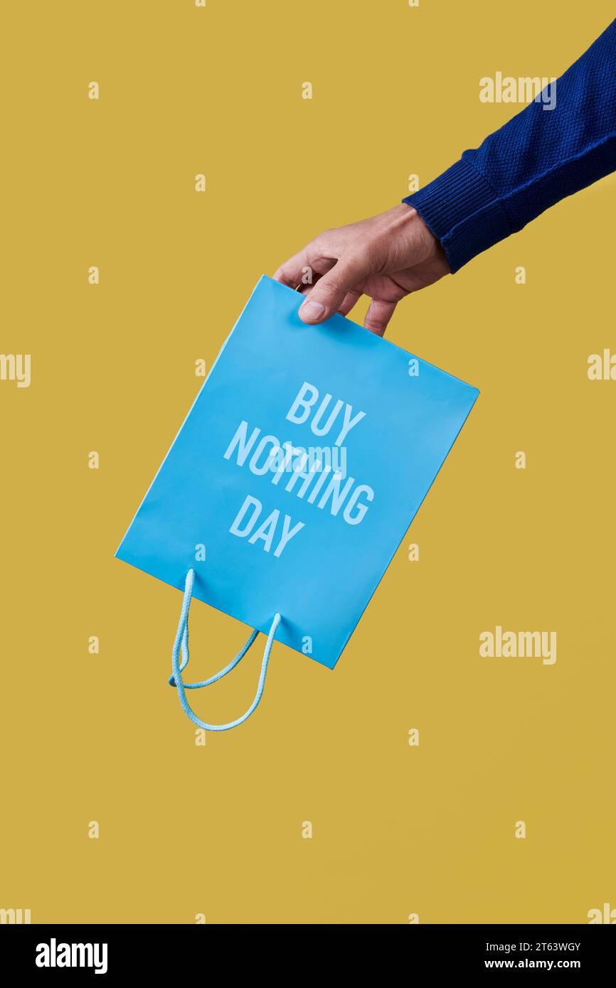 a man has a blue shopping paper bag, with the text buy nothing day written in it, upside-down in his hand, on a yellow background Stock Photo