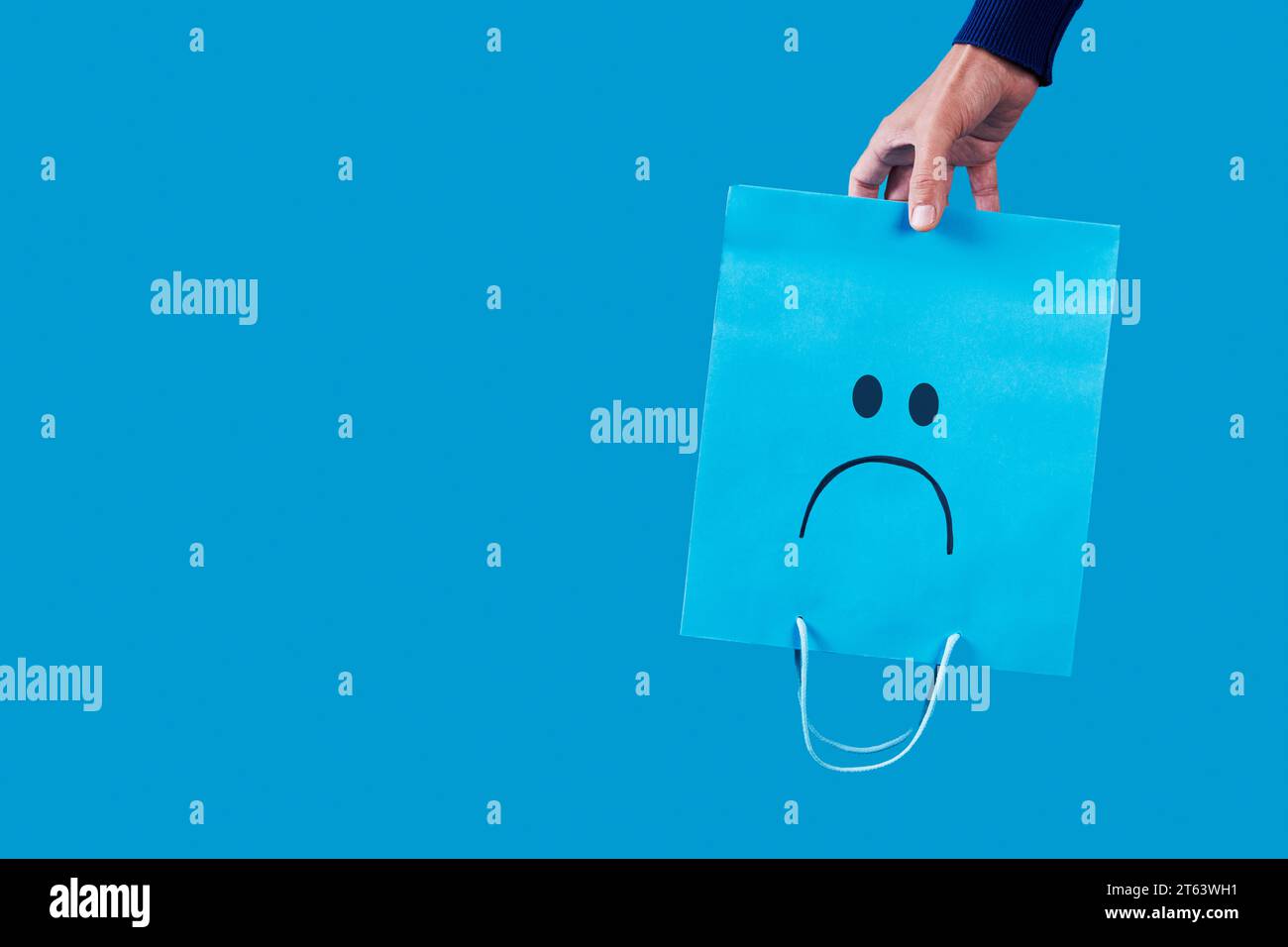 a man holds a blue shopping paper bag, with a sad face painted in it, empty upside-down on a blue background, with some blank space on the left Stock Photo