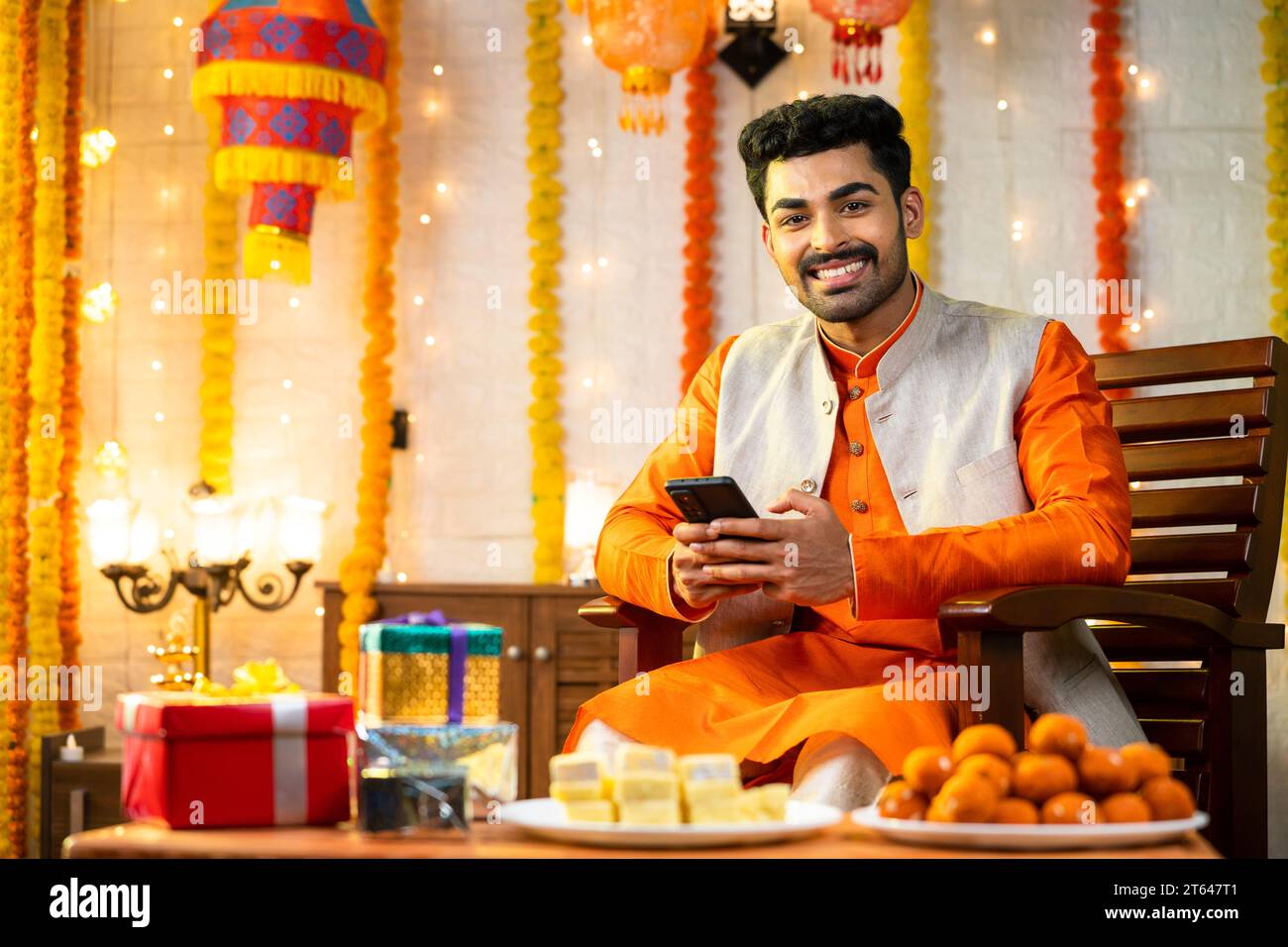 Happy smiling indian man looking at camera while using mobile phone on chair at home during diwali - concept of festival offers, enjoyment and Stock Photo