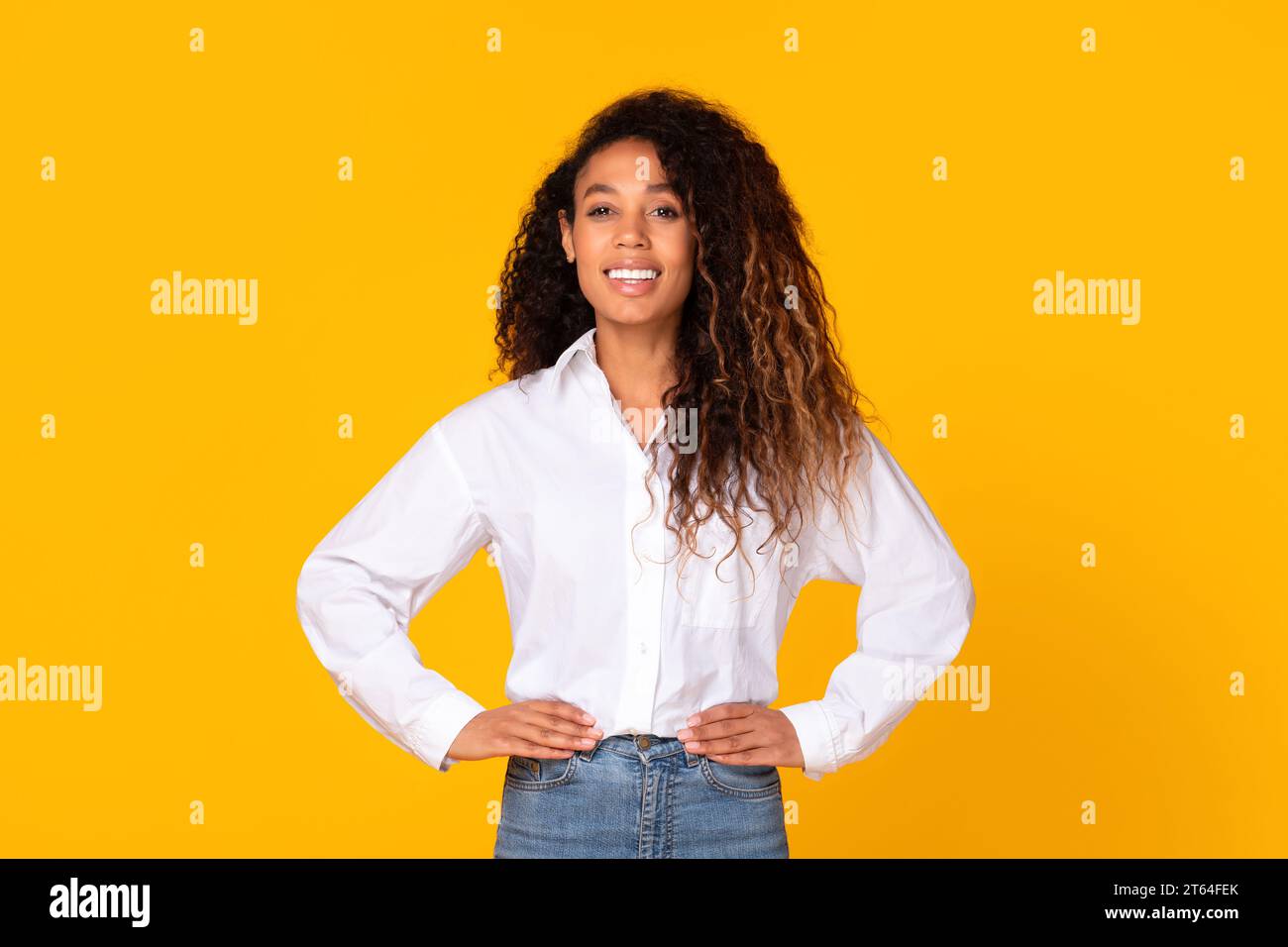 Black millennial lady standing holding hands on hips, yellow backdrop Stock Photo