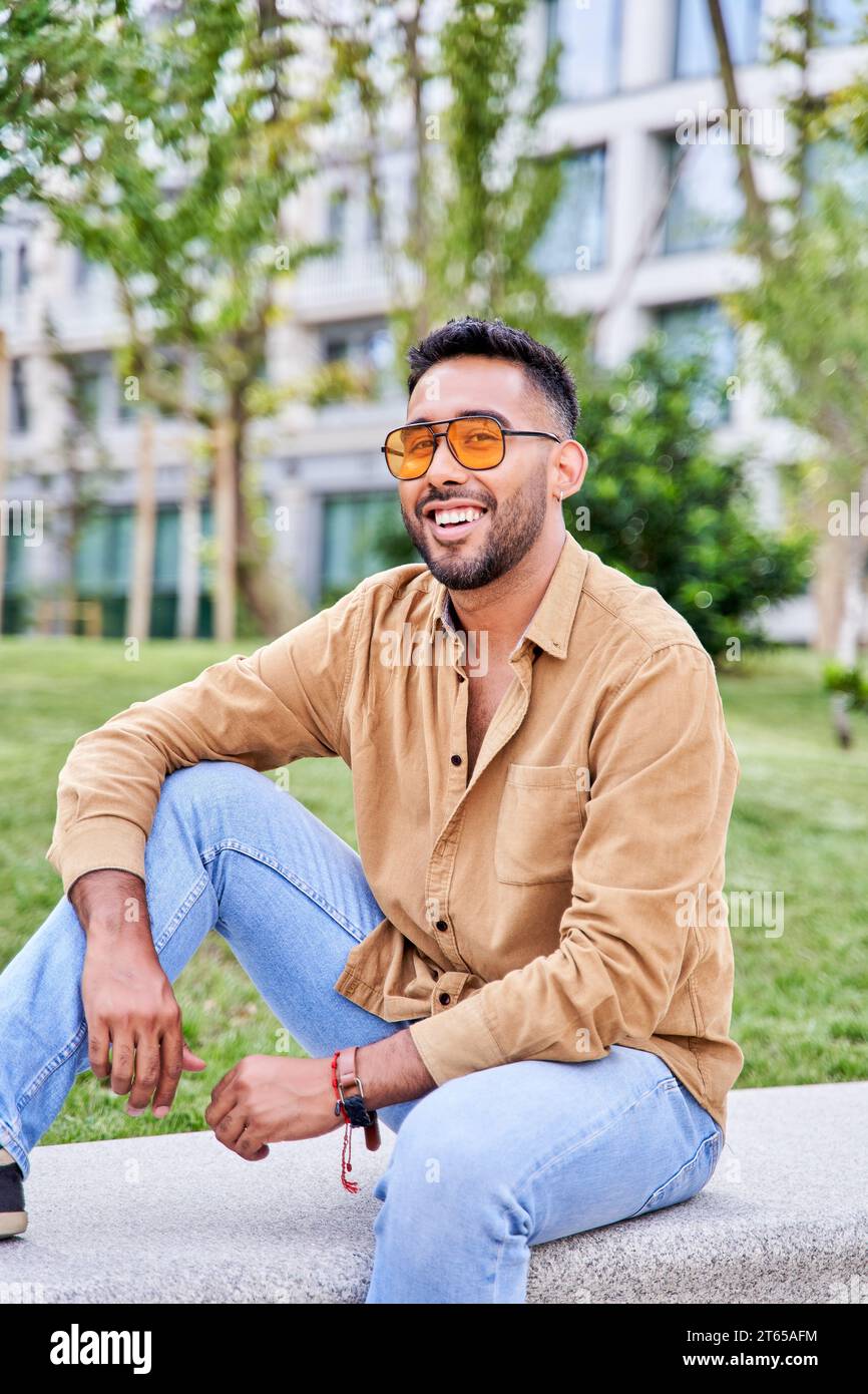 Young happy Hispanic man thinking while sitting at park. latin man wearing sunglasses Stock Photo