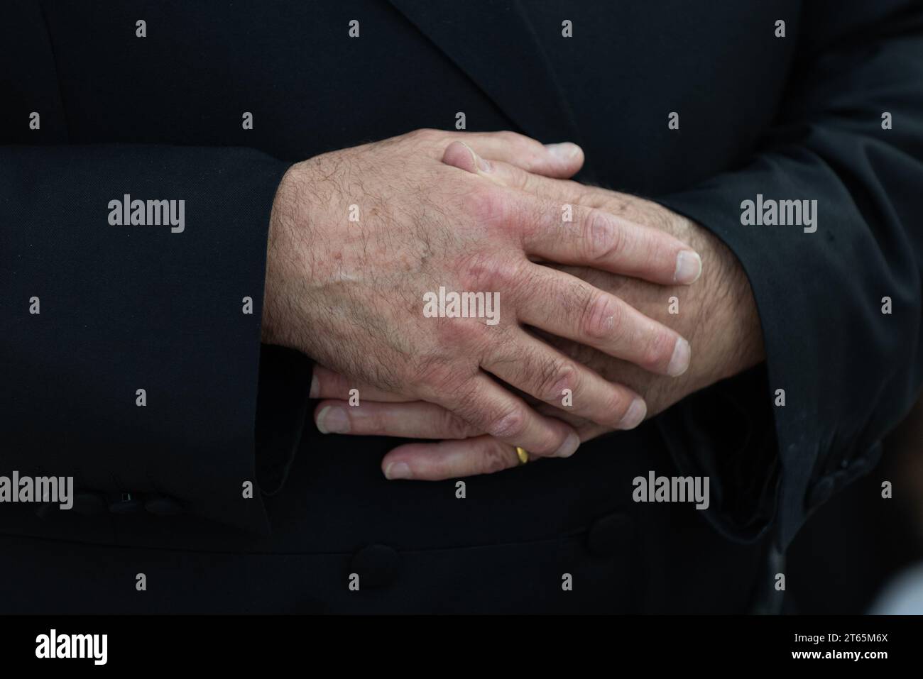 A man enfolds his hands and rests them on his body clothed in a long, black coat with buttons. Stock Photo