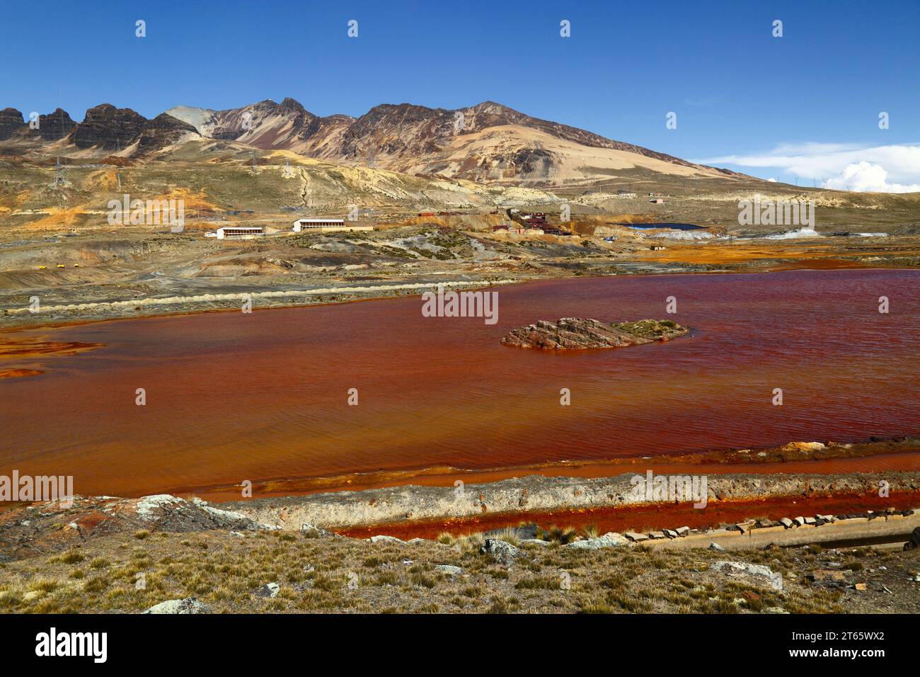 View across lake and aqueduct contaminated by acid mine drainage and industrial waste from the nearby tin mine at Milluni (part of which can be seen on the far bank and mountainside), near La Paz, Bolivia. The mountains are the foothills of the Mt Chacaltaya massif. Stock Photo
