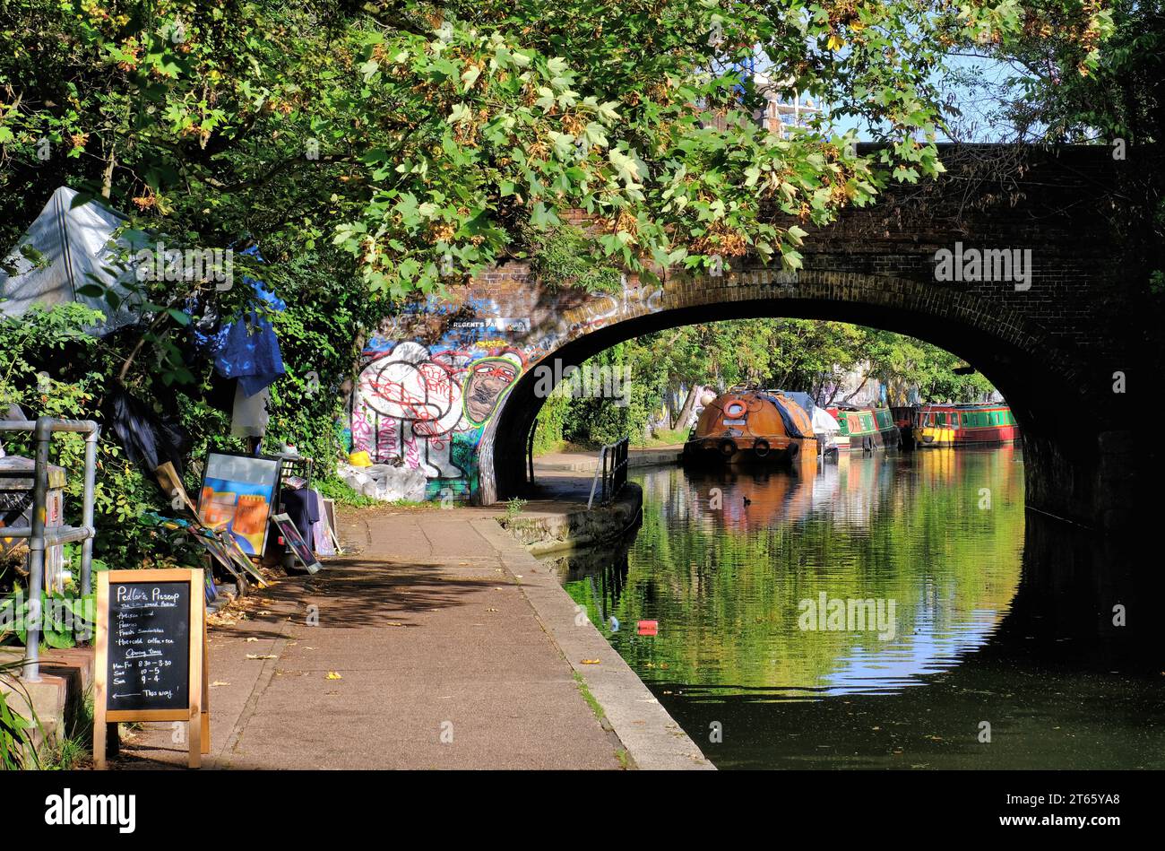 Arched brick bridge over Regents Canal at Regents Park Road with colourful graffiti, canal boats and reflections in Camden Town, London, England Stock Photo