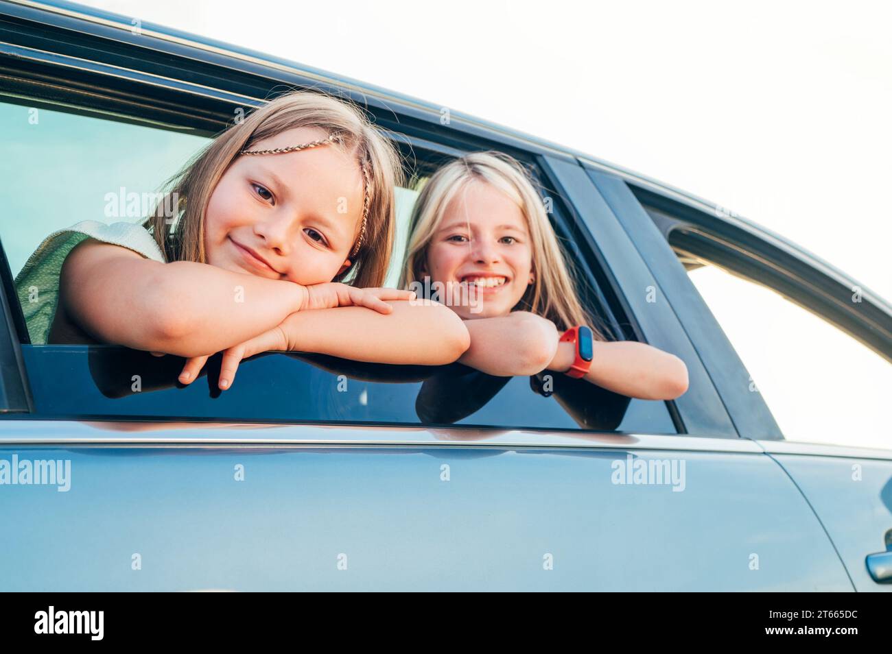 Two happy sisters looking out open car window during auto trip. Cute girls are smiling, laughing during road jorney. Family values, traveling concept Stock Photo
