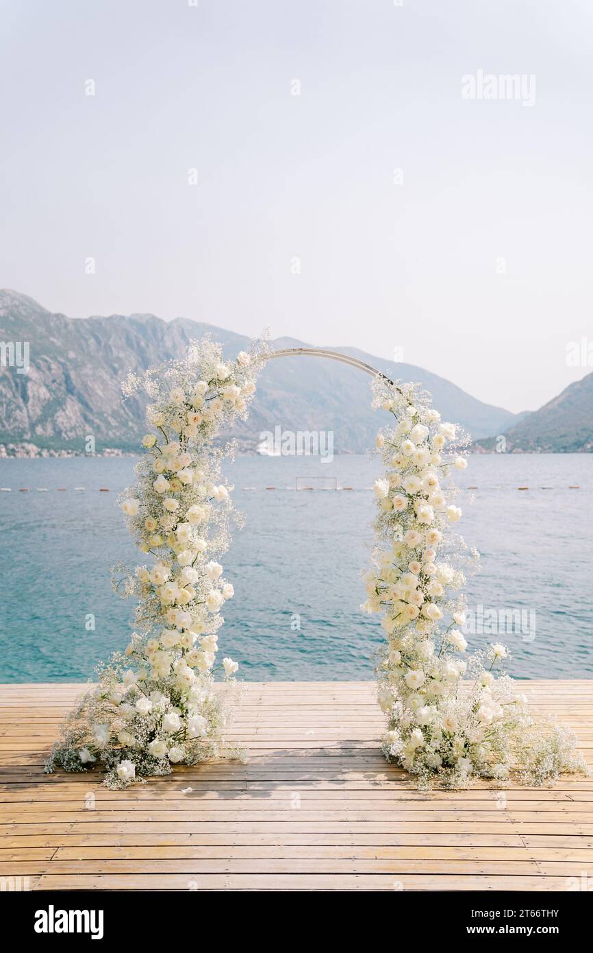 Wedding arch entwined with white flowers stands on a wooden pier by the sea Stock Photo