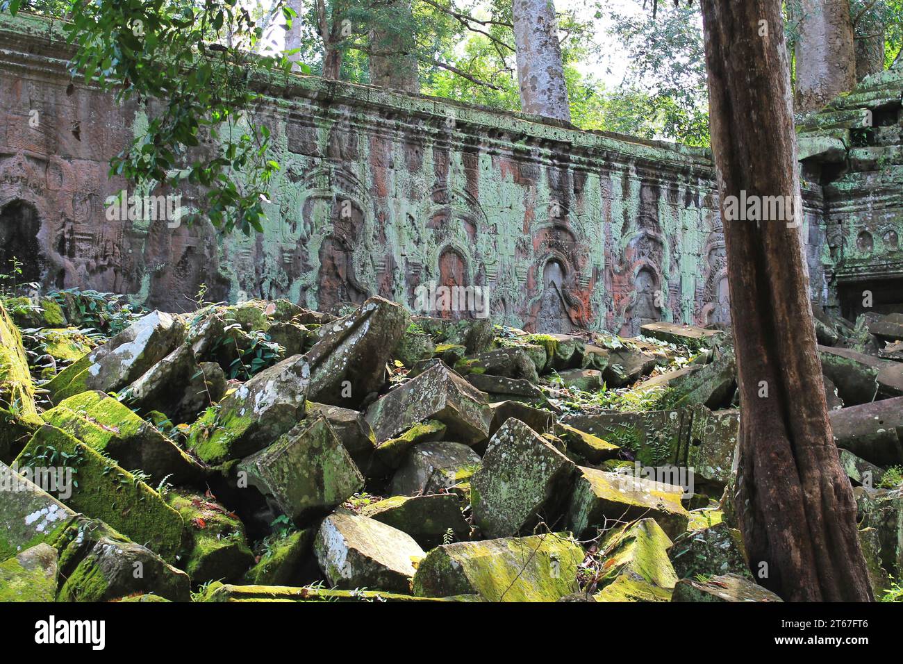 Mossy carved stone blocks in foreground with still standing ancient temple wall behind at the Angkor Archaeological Park near Siem Reap, Cambodia Stock Photo