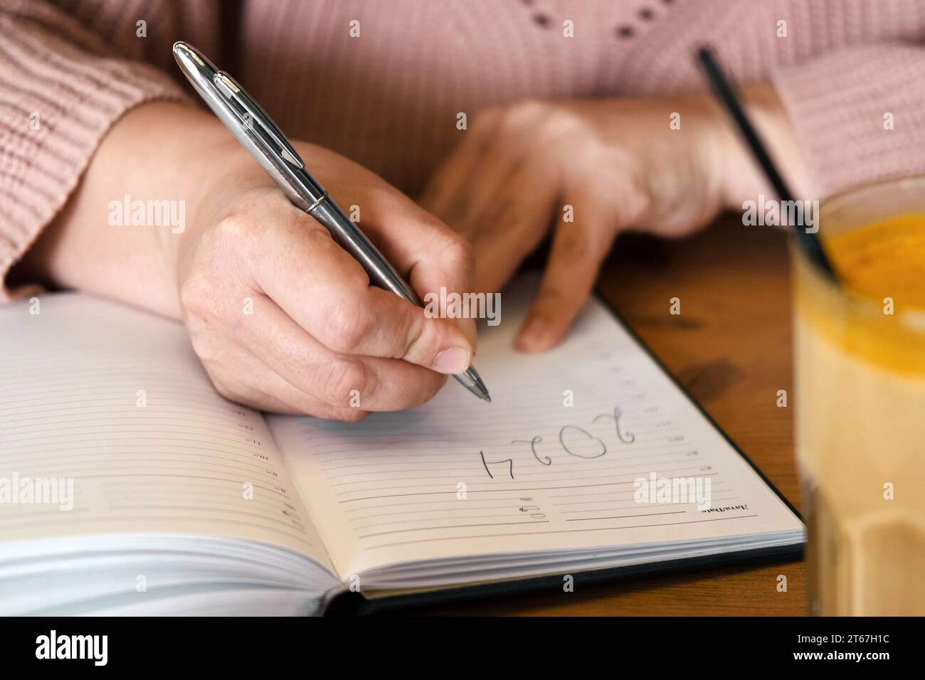 Woman's hand writing 2024. New Year Routine, resolutions on desk. Cafe, restaurant interior background. 2024 goals list with notebook, coffee cup on w Stock Photo