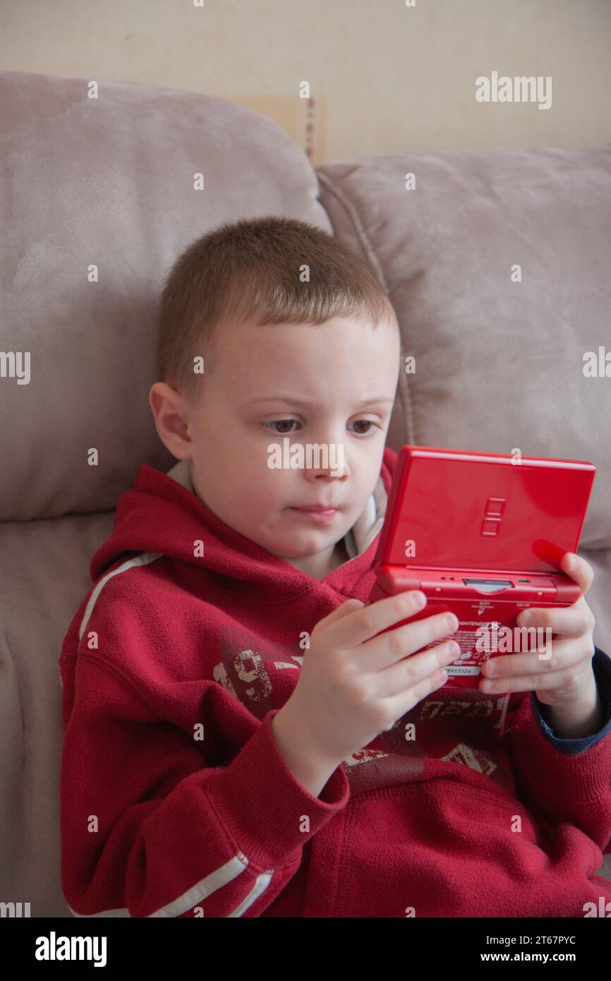 Young boy wearing a bright red hoody top sitting on a beige sofa and playing with a red handheld games console Stock Photo