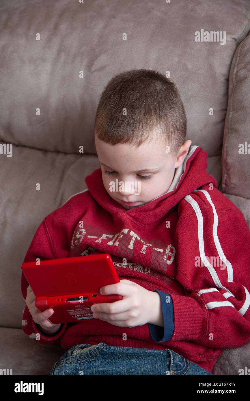 Young boy wearing a bright red hoody top sitting on a beige sofa and playing with a red handheld games console Stock Photo