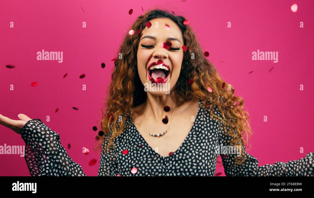 Beautiful young woman dances while red confetti glitter rains down, pink studio Stock Photo