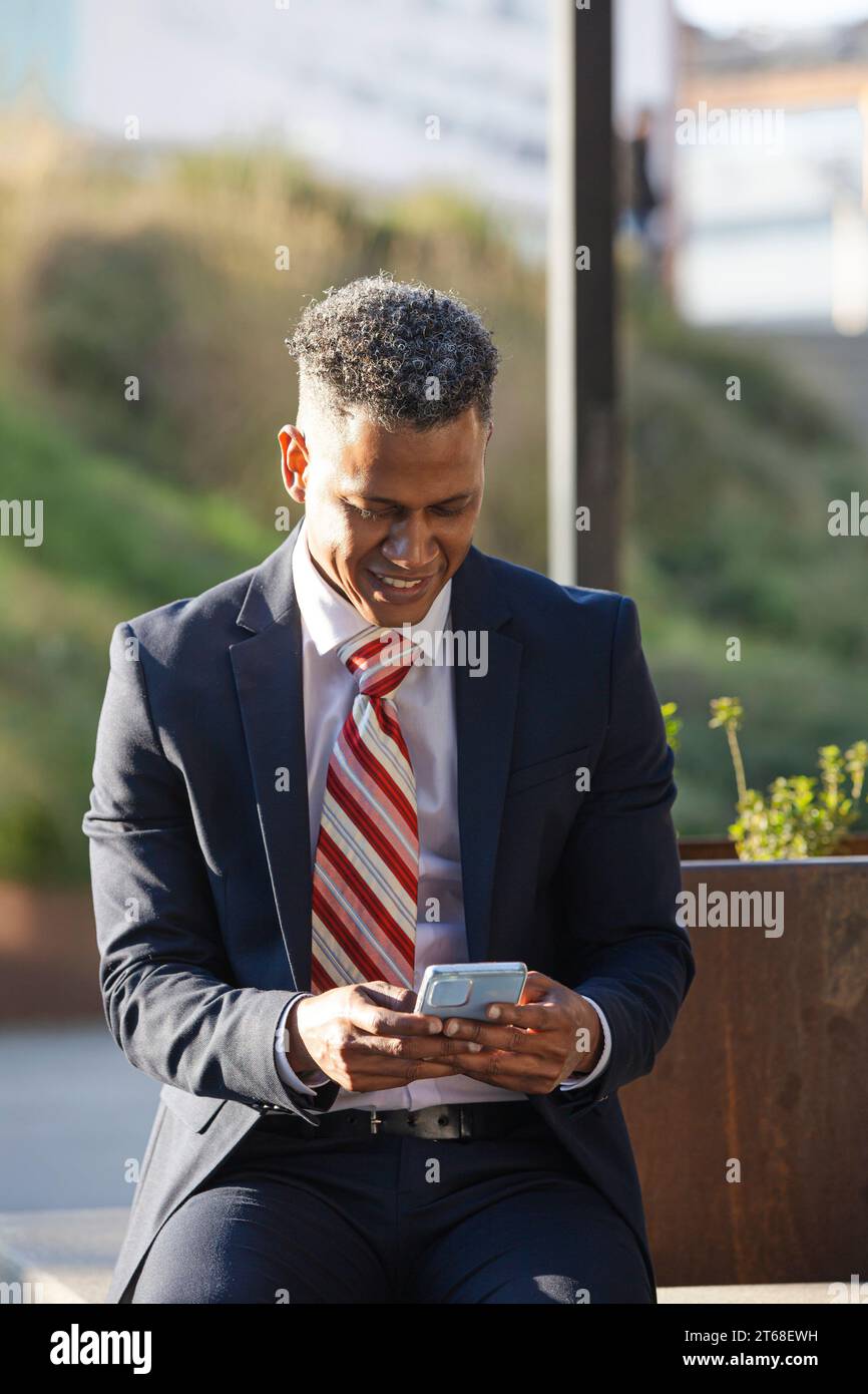 A young professional man in a suit sits in a park, using his phone for business communication Stock Photo