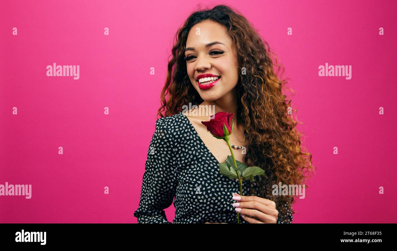 Beautiful young woman flirts with single stem rose, Valentine's Day, pink studio Stock Photo