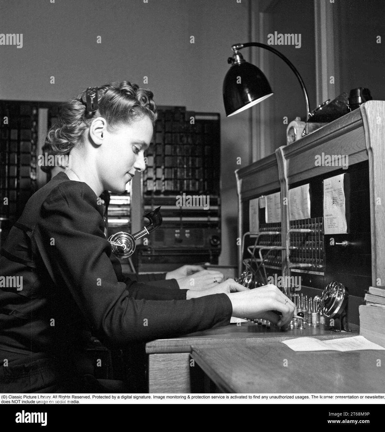 A young woman works in the company's switchboard 1942. She connects the incoming and outgoing calls to the right person using cords for the respective telephones in a so-called cord switch. The hairstyle is typical of the 1940s.  Sweden in 1942. Kristoffersson ref A120-1 Stock Photo