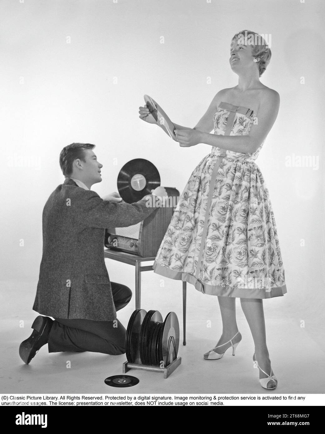 Gramophone in the 1950s. An man is kneeling by a combined recieving set and gramophone cabinet where he has opened the cover of the gramophone and holds a record in his hand.She is dressed in a typical 1950s dress looking happy.  Sweden 1959. Kristoffersson ref CD65-3 Stock Photo