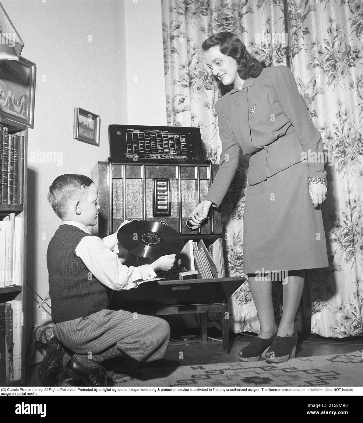 A young woman and her son at a radio gramophone with the option of playing gramophone records as well as listening to foreign radio channels via longwave, the cities of the various stations are written on the radio's panel. The boy is holding a 78-turn record of Bolero by Maurice Ravel. Sweden 1945. Kristoffersson ref M131-6 Stock Photo