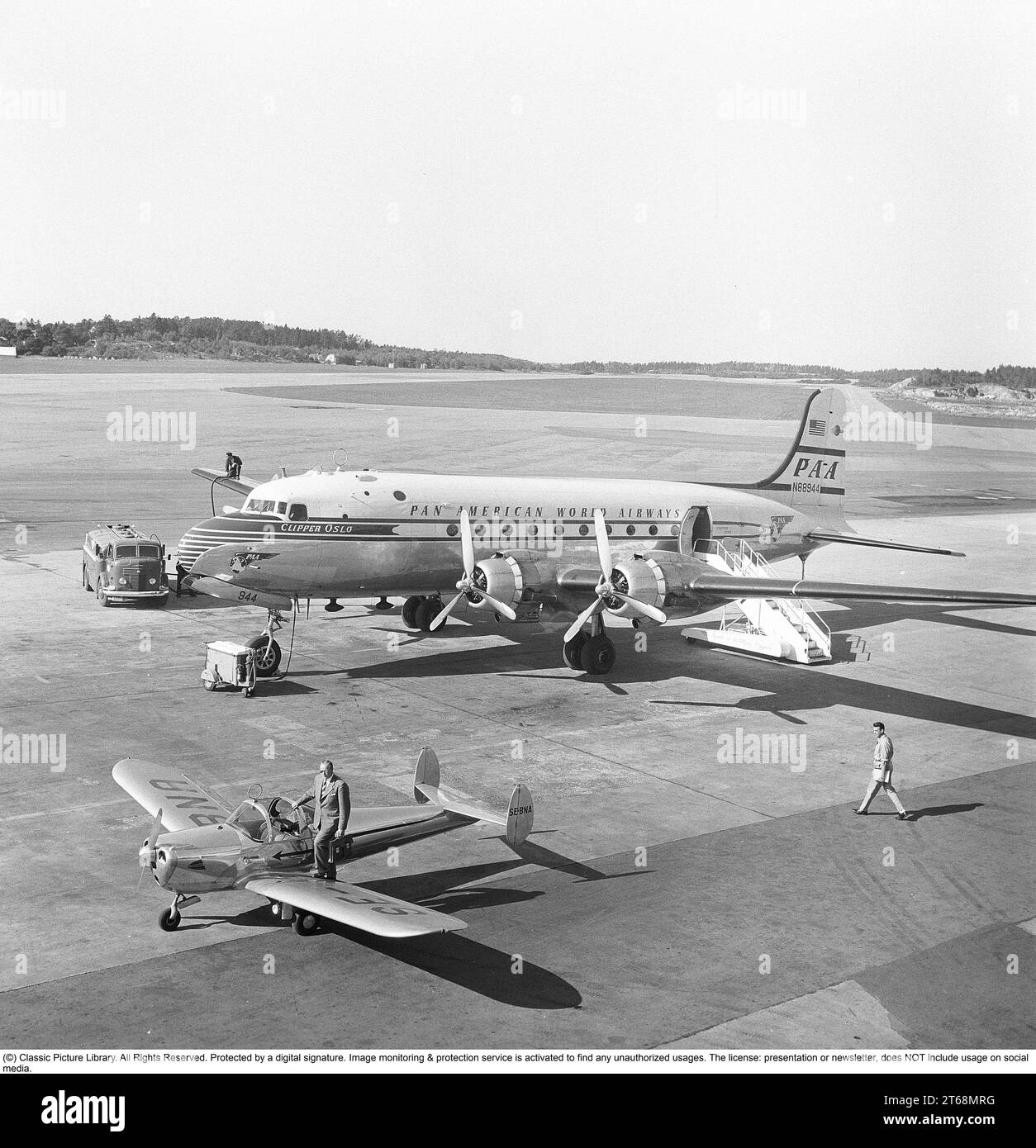 1950s airport. A Douglas DC-4 of Pan American World Airways 'Clipper Oslo' PA-A N88944 being prepared and fueld before takeoff. A man is seen on the wing at the end of the gas pipe from the truck.  The smaller propeller aircraft is a Ercoupe 415 SE-BNA with a man with a briefcase about to step into the aircraft.    Sweden 1953. Kristoffersson ref BM42-3 Stock Photo